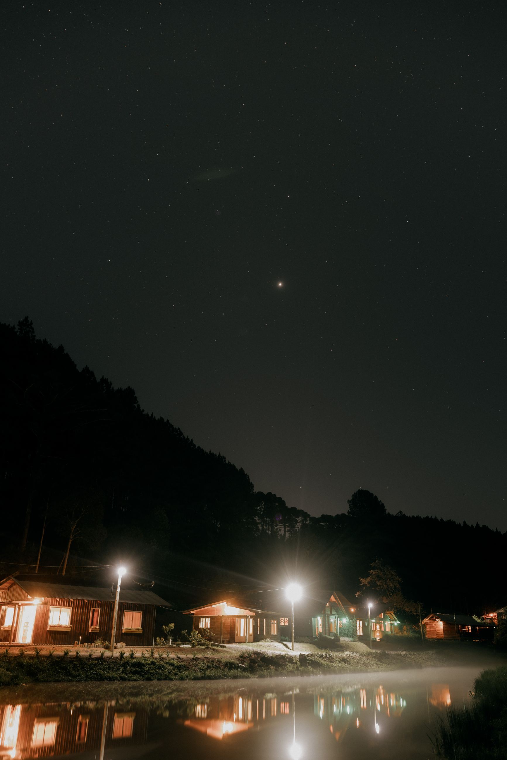 Cabanas iluminadas à noite, refletidas no lago sob um céu estrelado em Rio dos Cedros